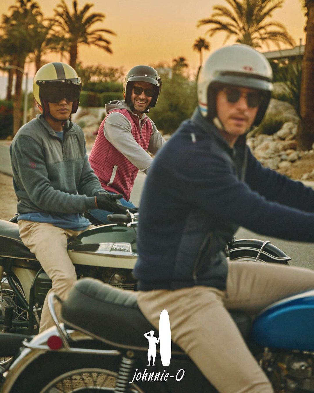 Three adult men riding motorcycles on a road lined with palm trees during a sunny day. They are wearing helmets and sunglasses.