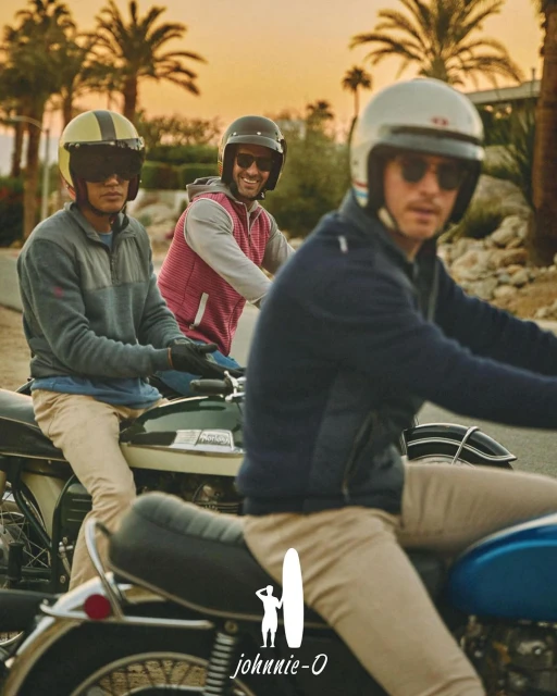 Three adult men riding motorcycles on a road lined with palm trees during a sunny day. They are wearing helmets and sunglasses.