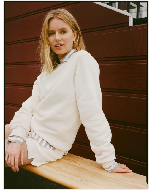Young woman in cream sweater and shorts sits on wooden bench, smiling. Dark red wall provides a simple studio background. Soft daylight.
