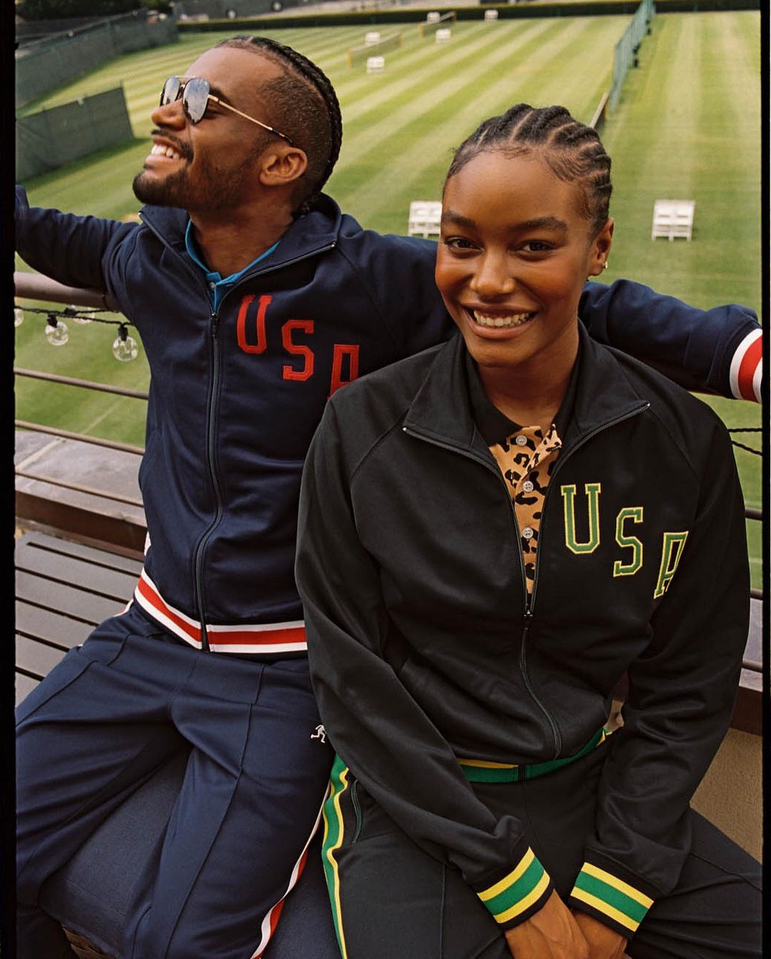 Two smiling athletes in USA tracksuits pose outdoors against a tennis court. A man in sunglasses and a woman with braided hair are seen.