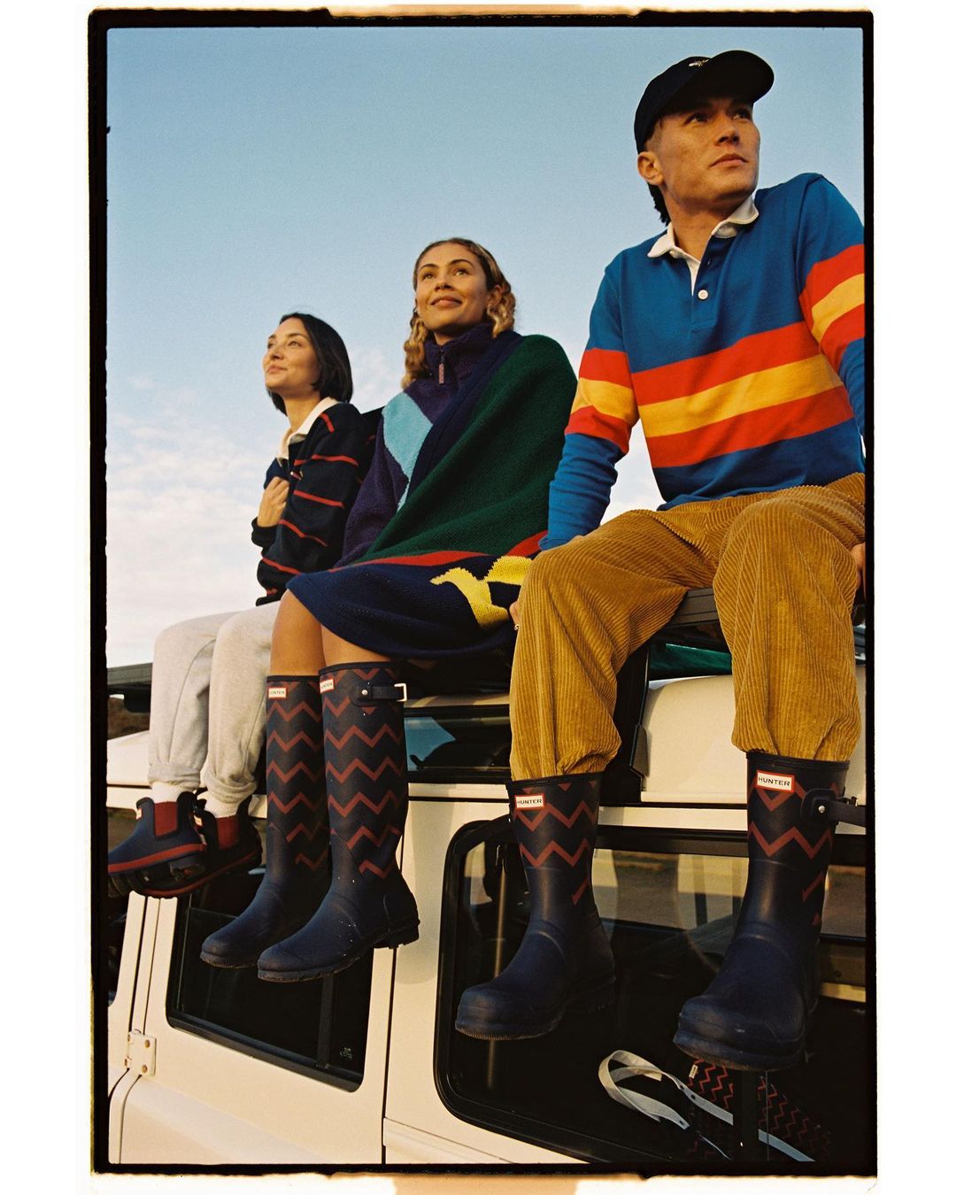 Three people sit on a white SUV, wearing colorful casual outfits and Hunter rain boots against a clear sky in an outdoor setting.