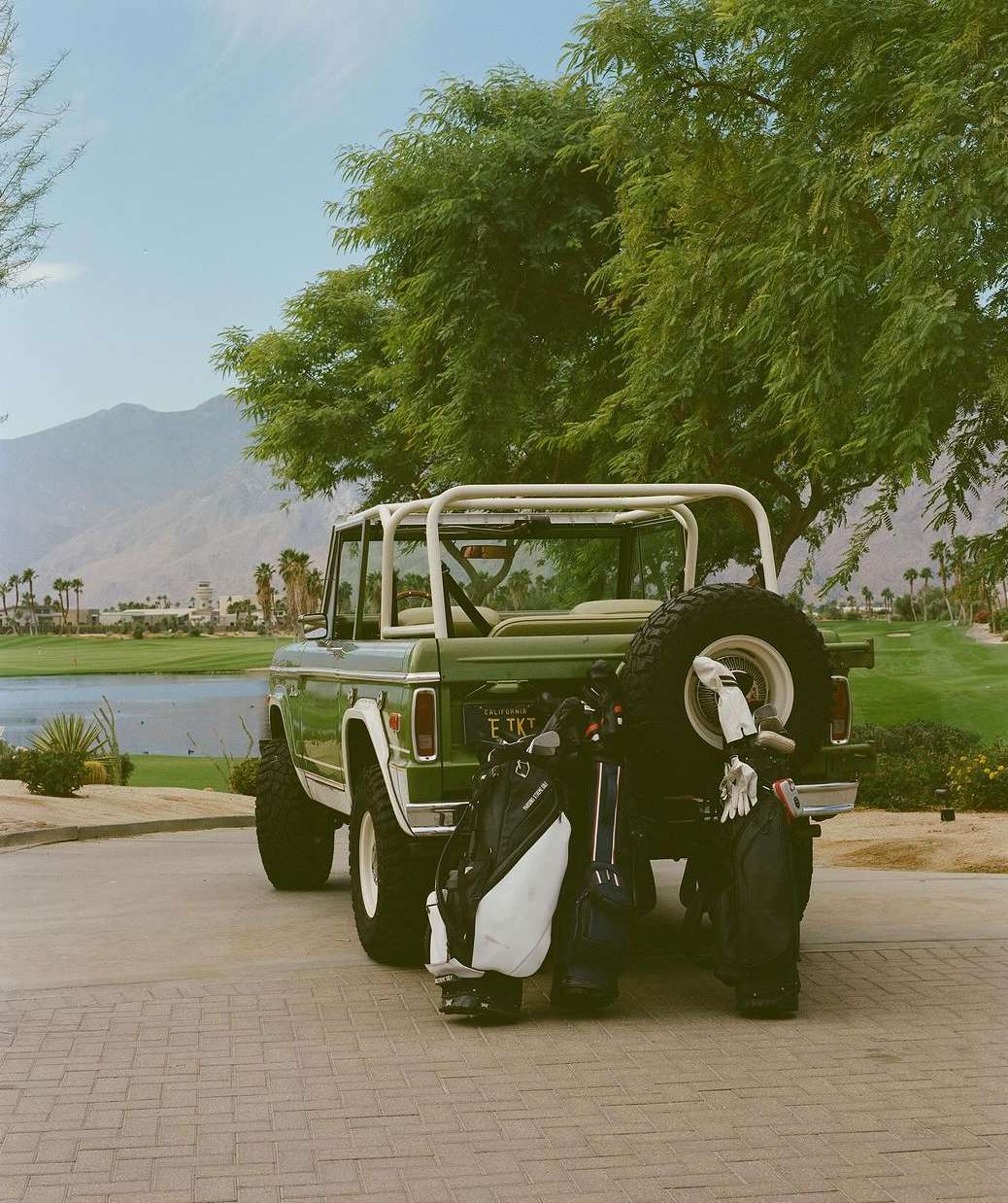 Green vintage Ford Bronco with golf bags parked on brick, set against a lush golf course and mountain backdrop on a sunny day.