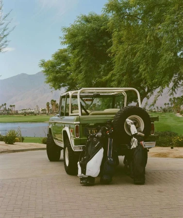 Green vintage Ford Bronco with golf bags parked on brick, set against a lush golf course and mountain backdrop on a sunny day.
