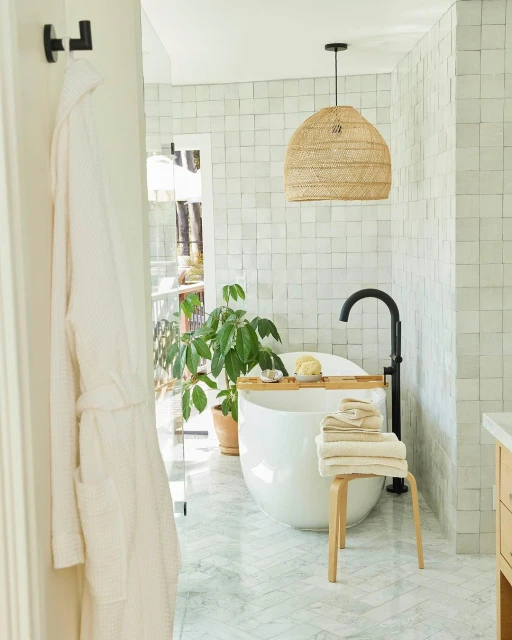 Bathroom interior with white freestanding tub, stool with towels, potted plant, straw pendant light, and white marble tile.