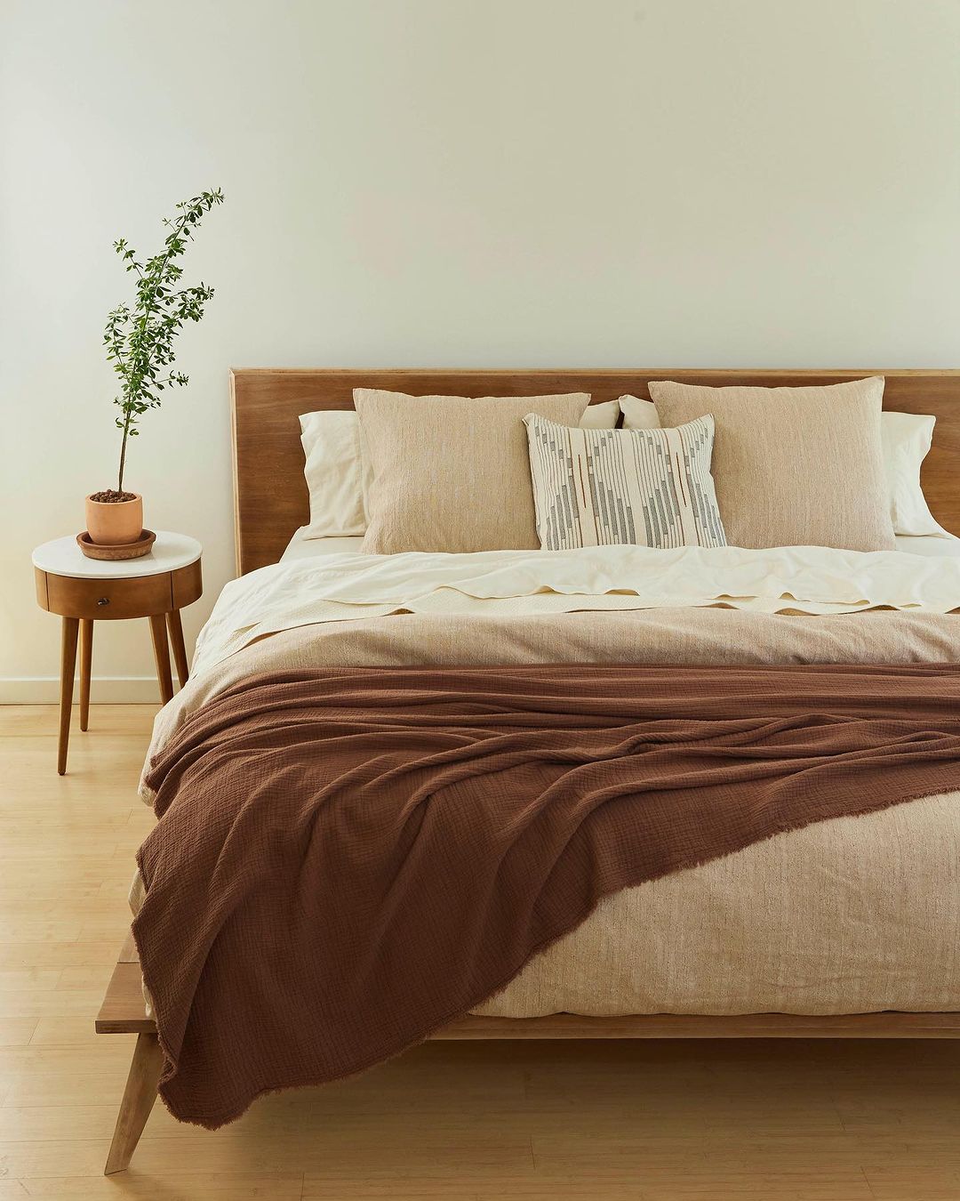 Interior of a minimal bedroom features a wooden platform bed with a brown knit blanket, neutral pillows, and a small plant on a side table.