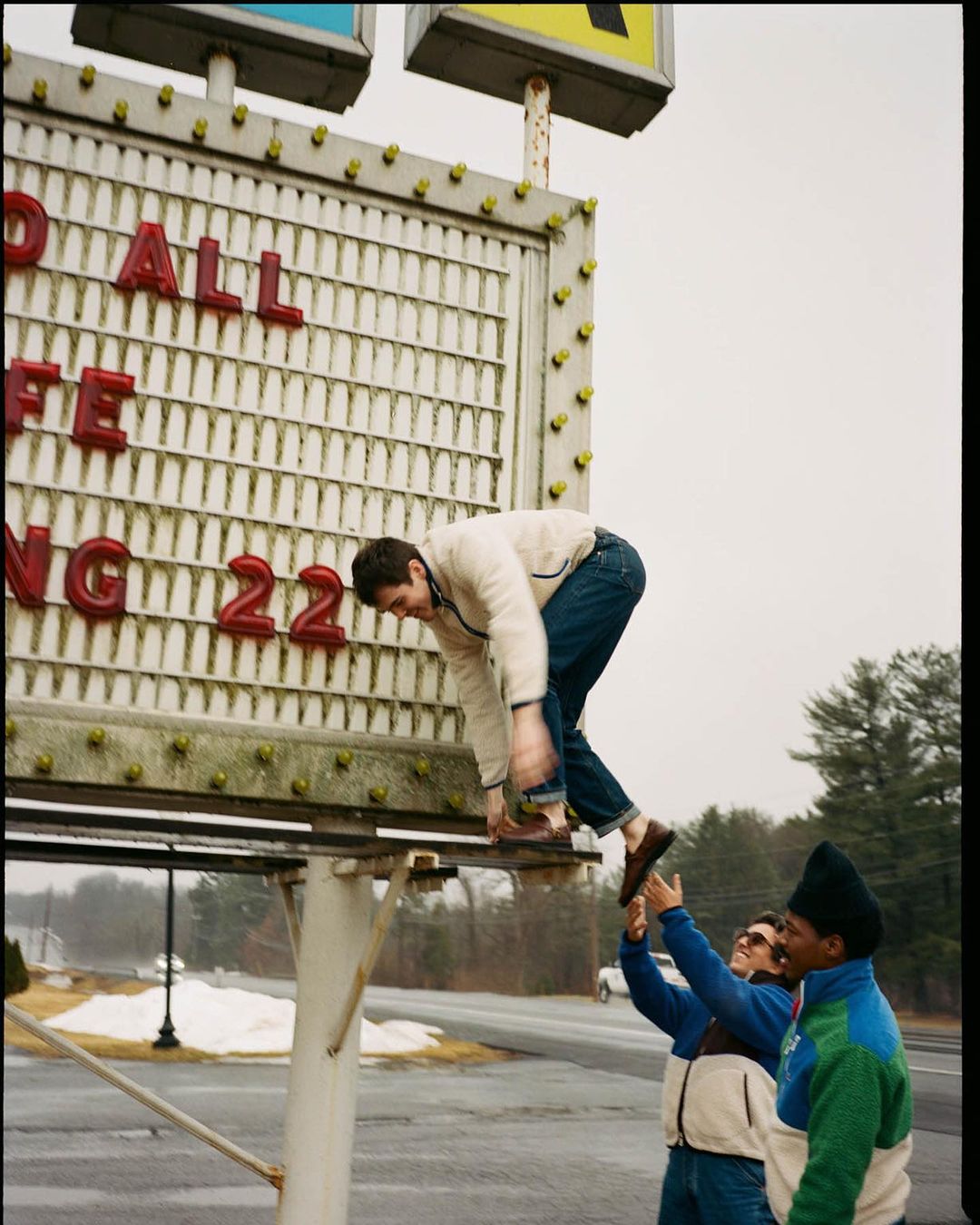 Man climbing up the side of a large sign as two other men help stabilize him near a road in a rural environment during daylight.