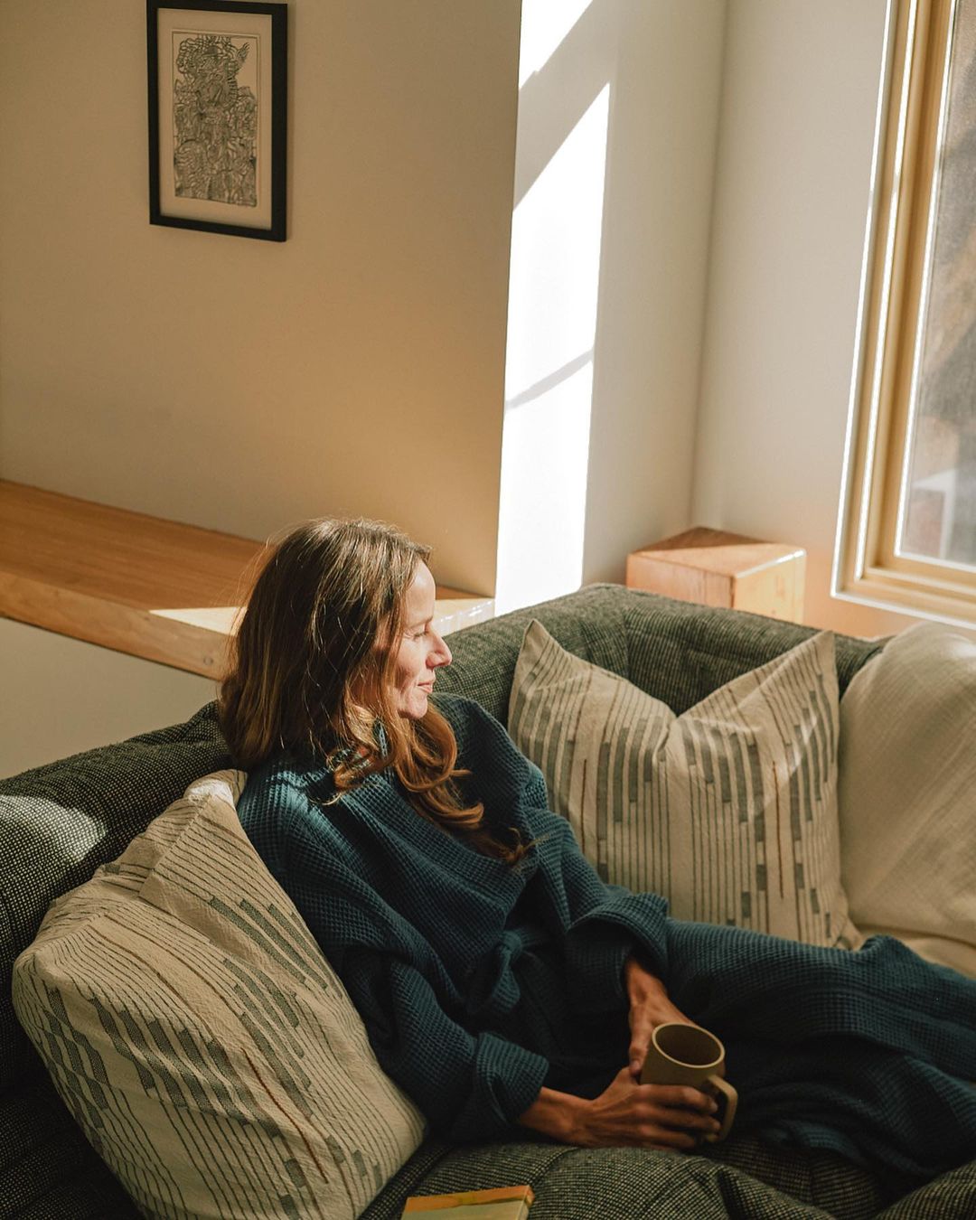 A woman in a blue robe relaxes on a sofa in a sunlit room, holding a mug near a window. Pillows and a book add to the cozy scene.