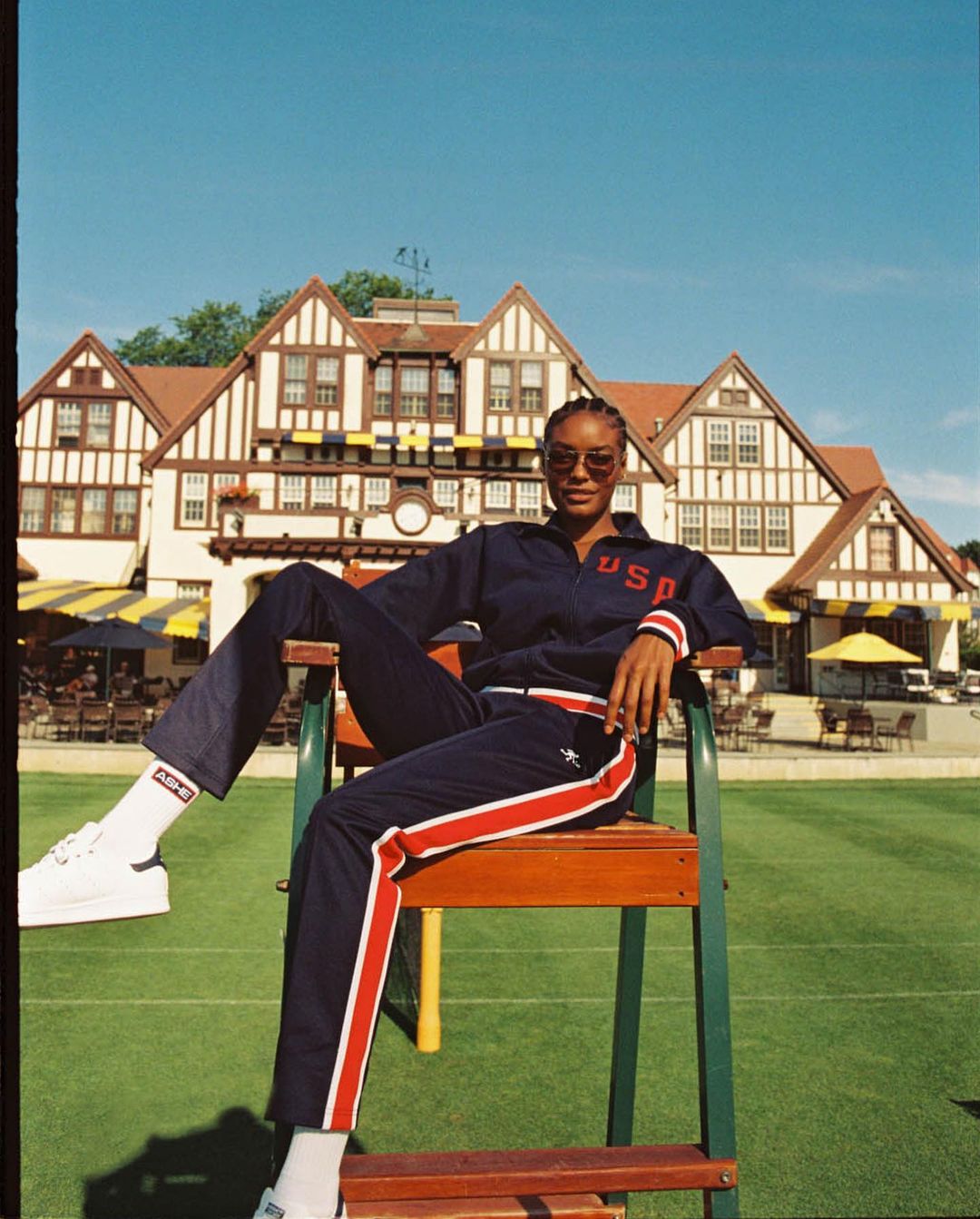 Woman in a USA tracksuit sits on a tennis umpire chair on a green tennis court with a red roofed clubhouse in the background on a sunny day.