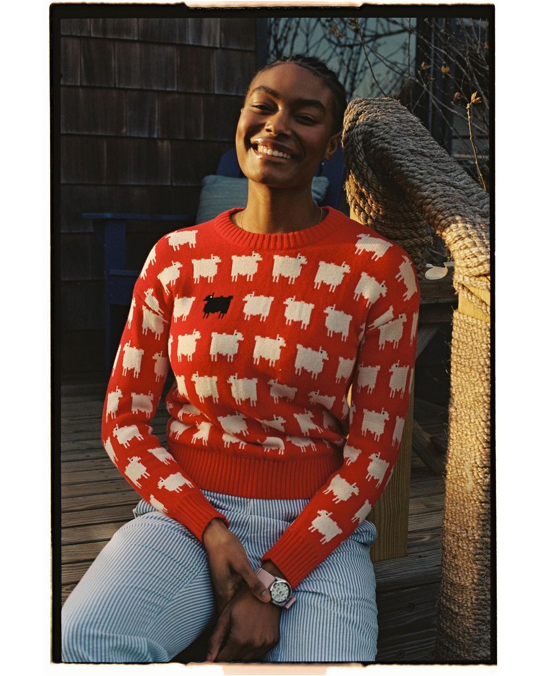 Smiling African American woman in a red sweater with sheep pattern poses outdoors on a wooden porch with a rope railing.