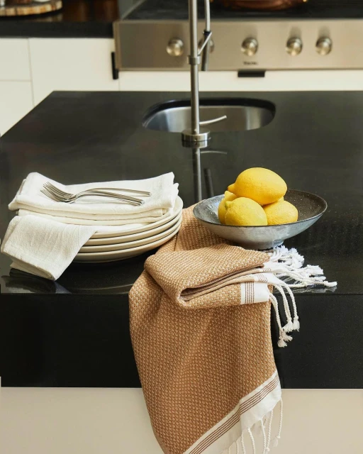Close-up shot of kitchen counter with plates, silverware, lemons in a bowl, and a textured towel, evoking a fresh and clean aesthetic.
