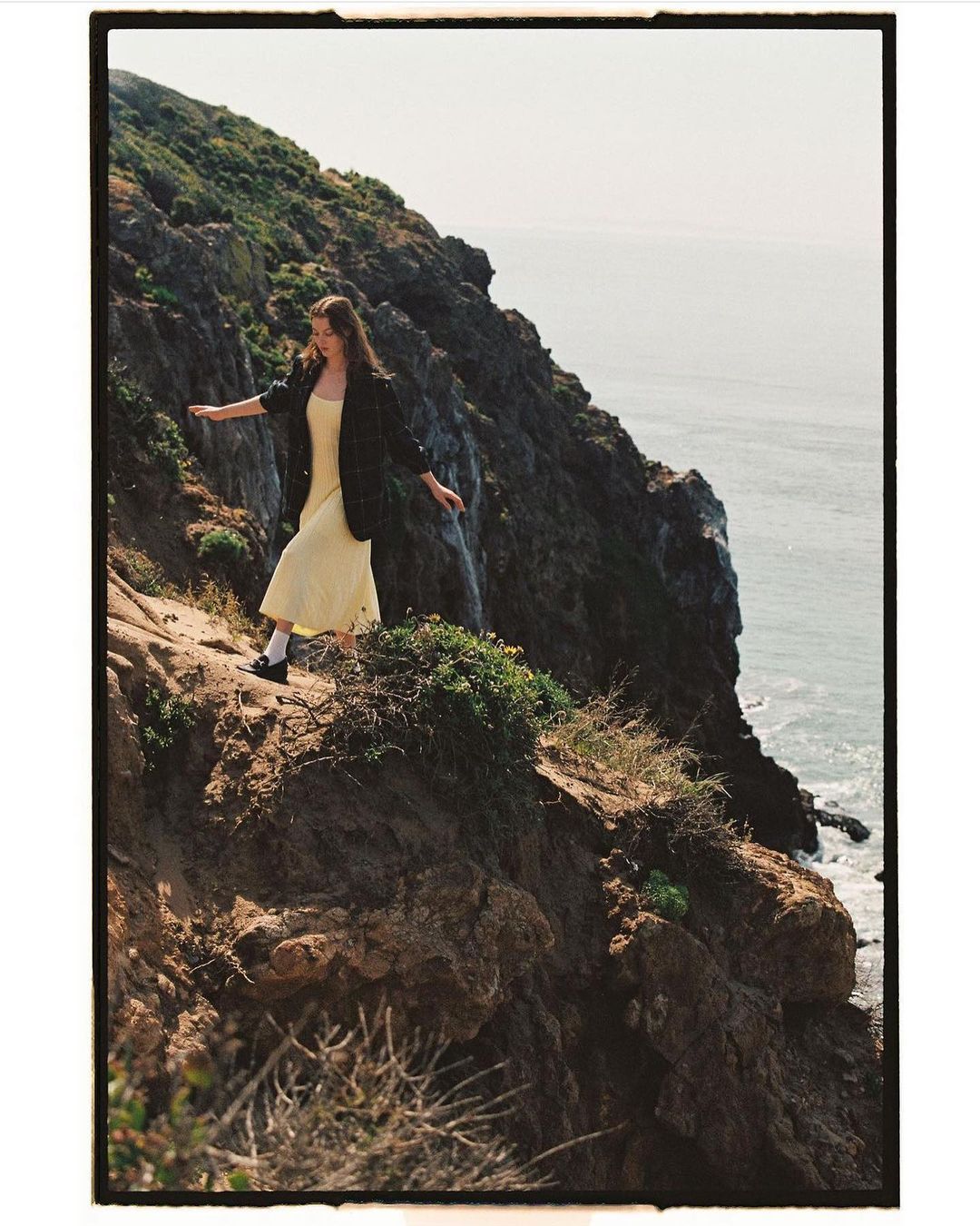 Model in yellow dress and dark blazer walking along a rugged rocky coastal cliffside with ocean views.