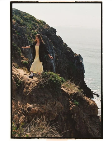 Model in yellow dress and dark blazer walking along a rugged rocky coastal cliffside with ocean views.