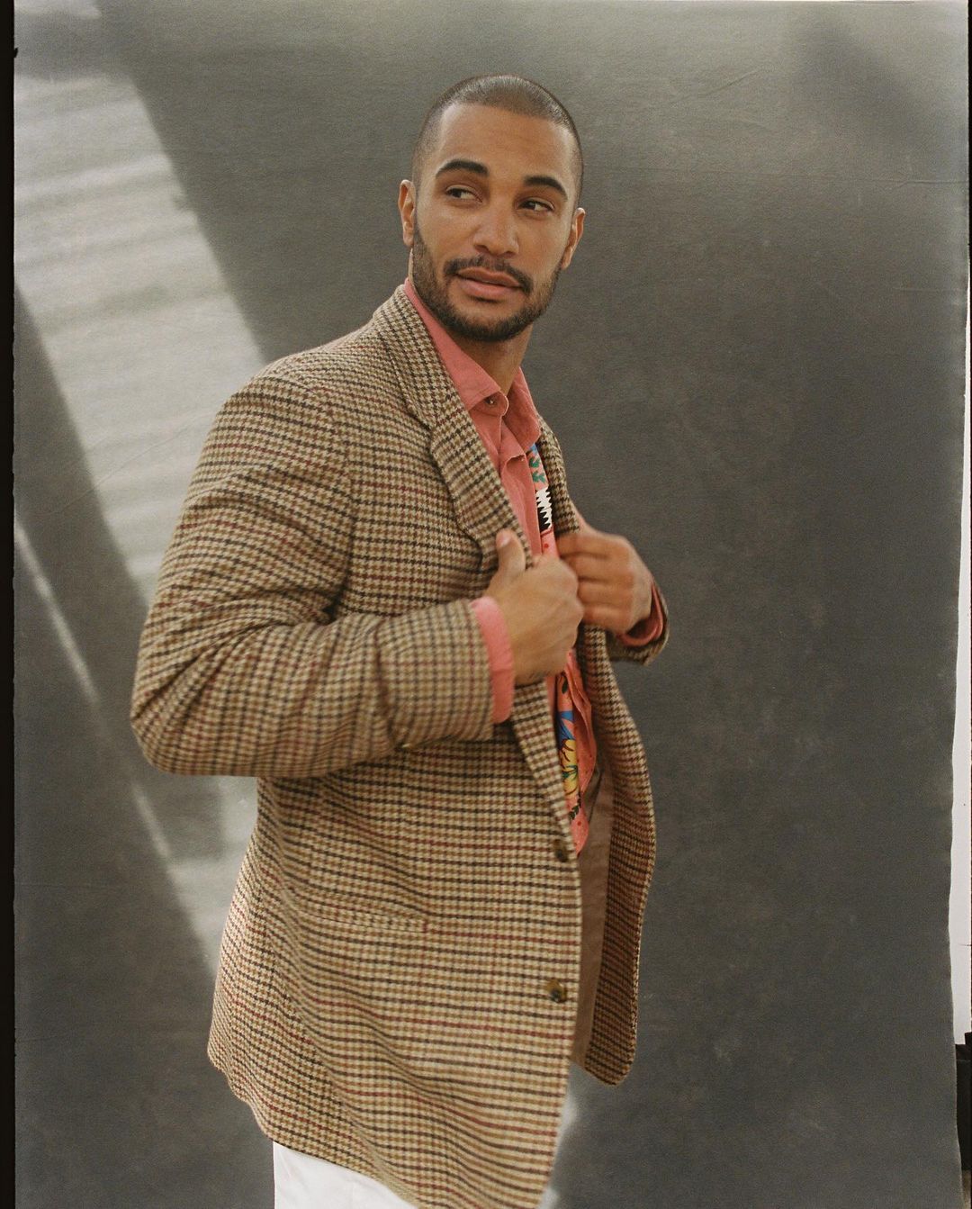 Portrait of an adult male in a checkered blazer over a salmon-colored shirt, looking over his shoulder in a studio setting.
