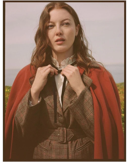 Woman in vintage-style plaid blazer and red cape poses outdoors. Portrait shot in natural light with soft focus, emphasizing a fashion aesthetic.