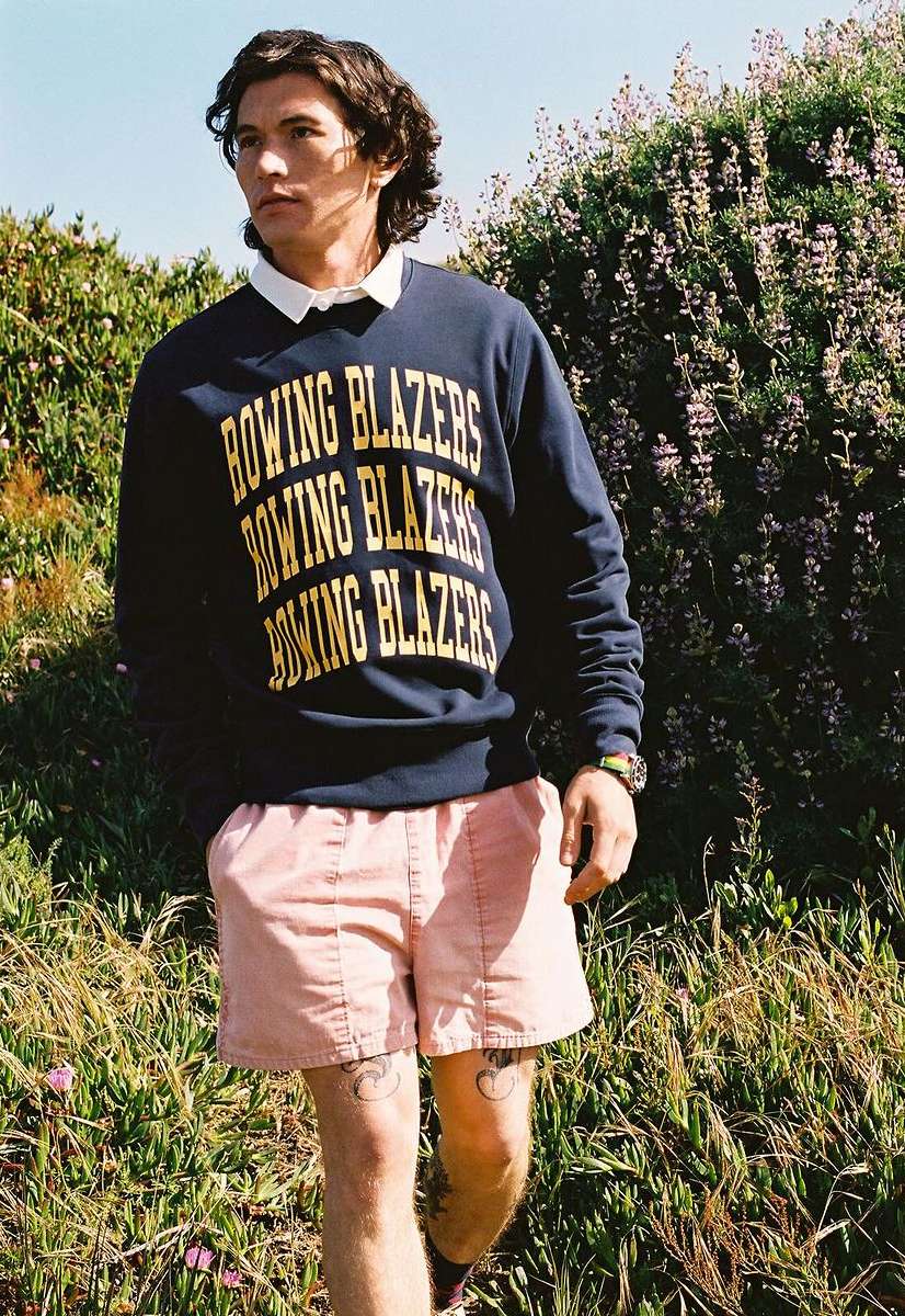 Man in Rowing Blazers sweatshirt and pink shorts stands in a grassy, outdoor setting, showcasing casual fashion and a relaxed style.