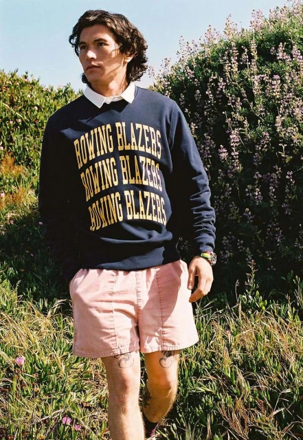 Man in Rowing Blazers sweatshirt and pink shorts stands in a grassy, outdoor setting, showcasing casual fashion and a relaxed style.