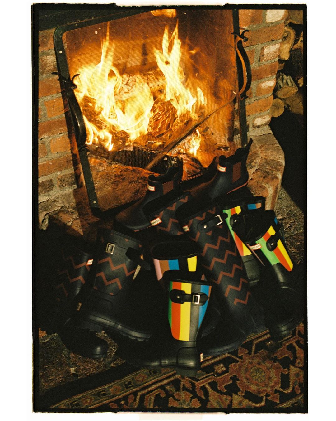 An arrangement of black rubber boots with colorful stripes and chevron patterns sits near a brick fireplace with a blazing fire.