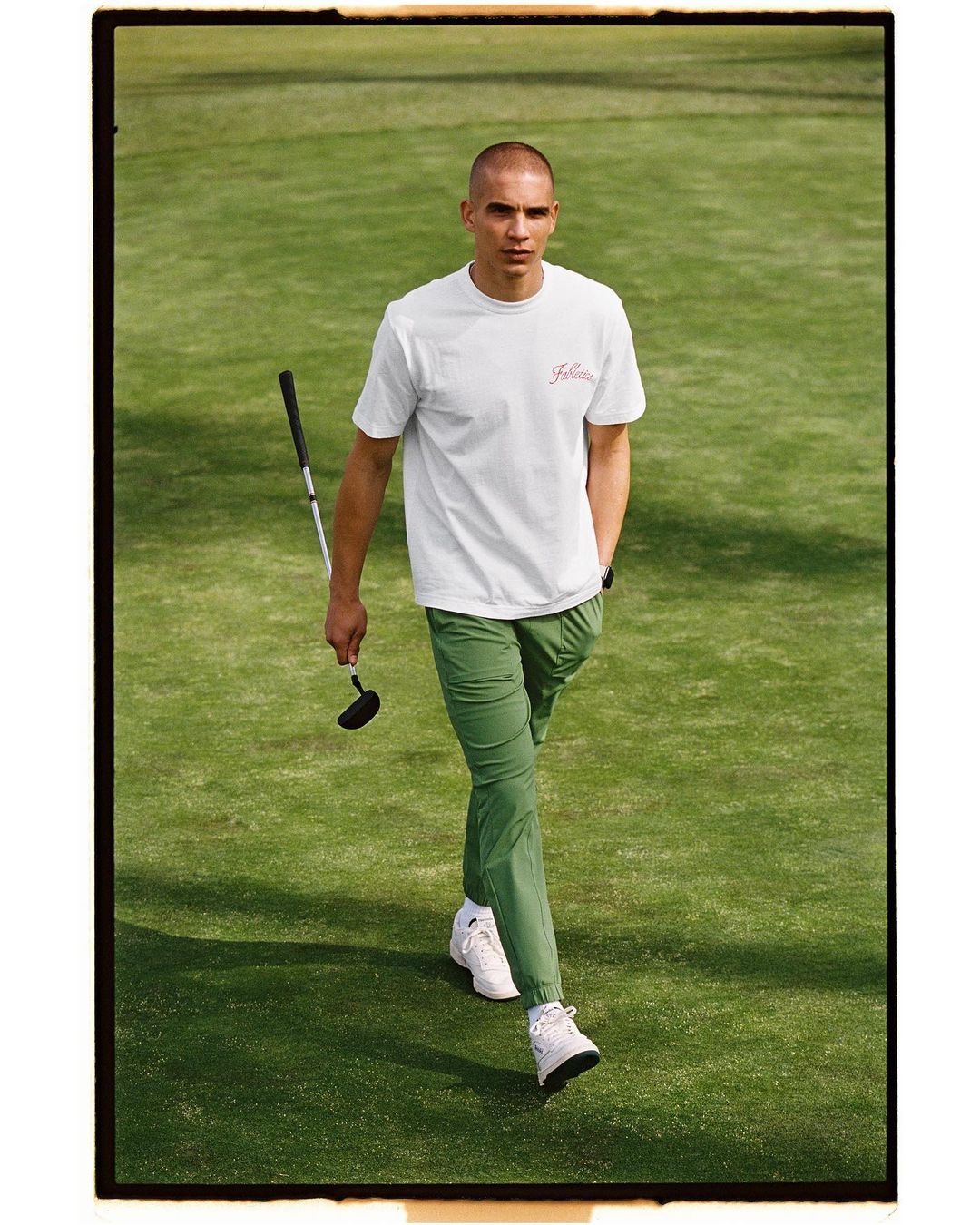 Athletic man in white tee and green pants walks on manicured green lawn holding golf club, full shot, outdoors.