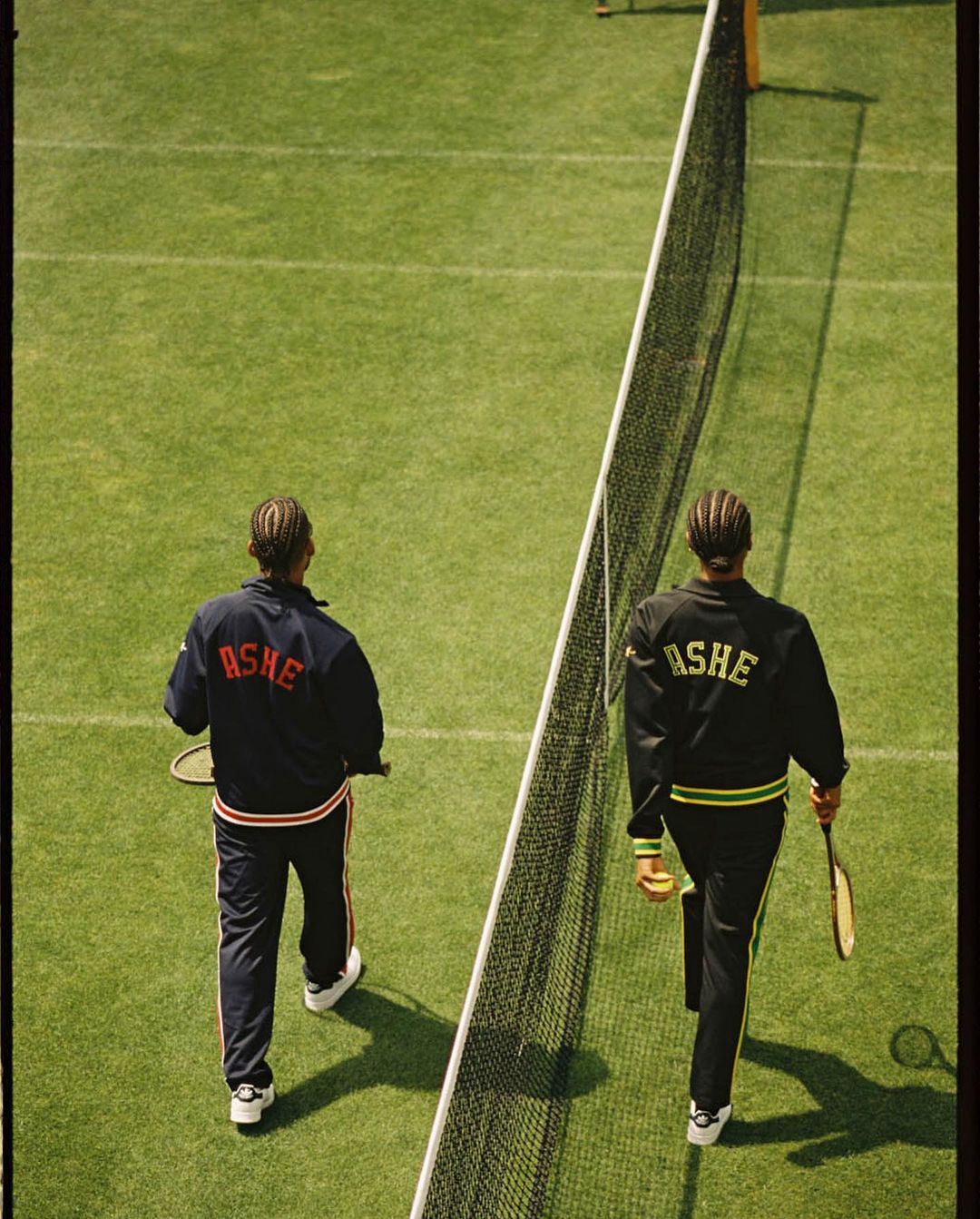 Two men in matching tracksuits walk onto a green tennis court with a net in the foreground, holding rackets and a tennis ball.