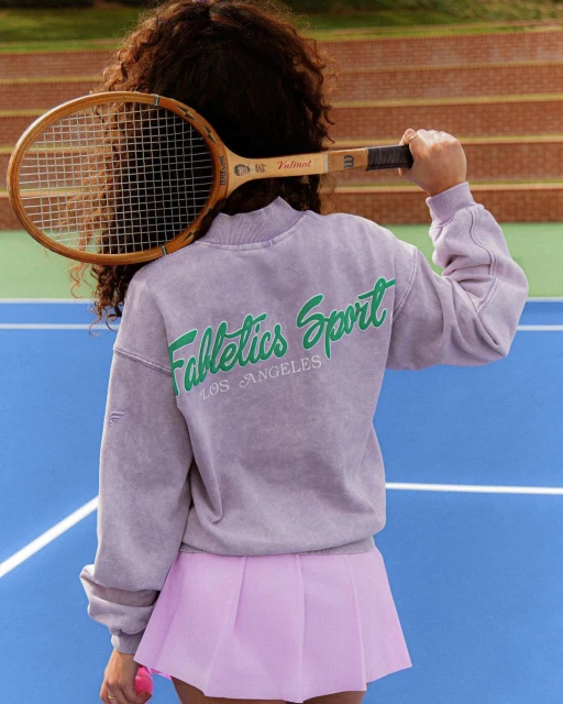 Woman with tennis racket on tennis court wearing sweatshirt with 'Fabletics Sport Los Angeles' on the back and a pink tennis skirt.