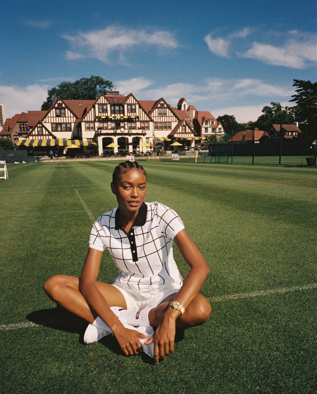 Woman sits cross-legged on grass in sportswear with a vintage building, blue sky, and white clouds in the background.