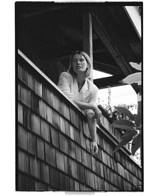 Black and white portrait of a woman in a light-colored blazer looking out from behind a wood shingle wall with greenery behind.