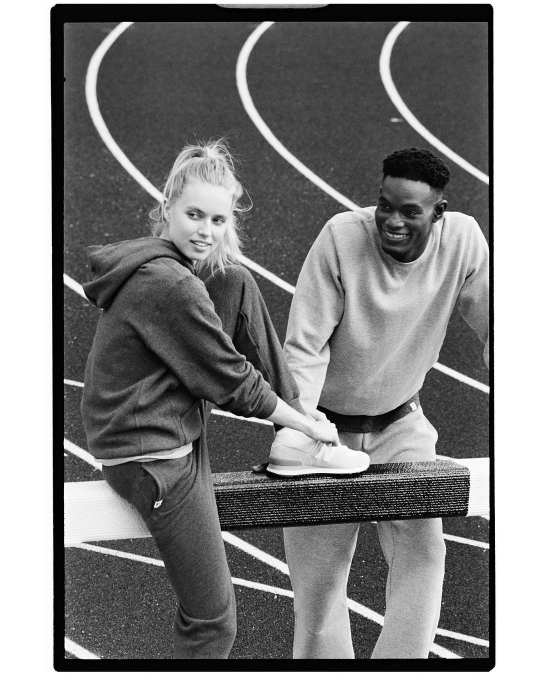 Two athletic people in sweatsuits pause for a break at a track and field venue, one with a foot up on a barrier, black and white.