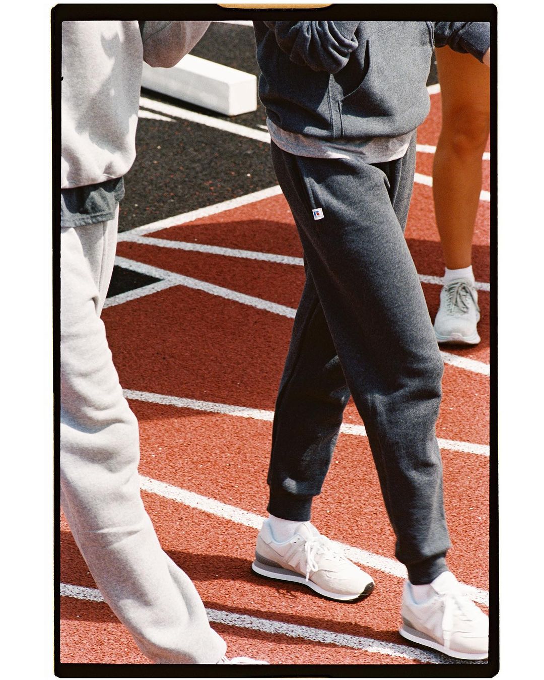 Three people walk on a red running track wearing grey sweatpants, hooded sweatshirts, and white sneakers in an outdoor setting.