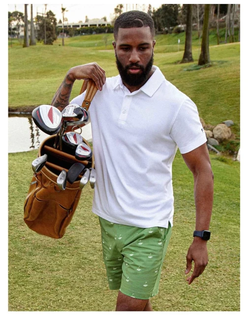 Adult male golfer poses outdoors with golf bag and golf clubs. He wears a white polo shirt and patterned green shorts. Lush golf course.