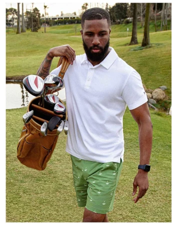 Adult male golfer poses outdoors with golf bag and golf clubs. He wears a white polo shirt and patterned green shorts. Lush golf course.