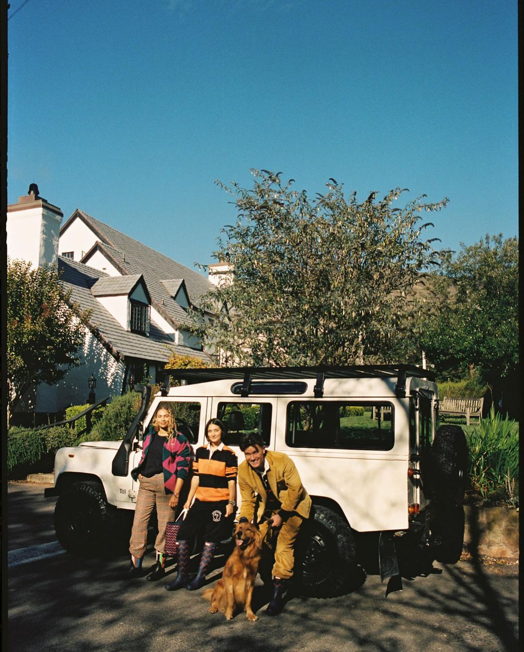 Three people pose with a golden retriever next to a white Land Rover SUV on a sunny day in a residential neighborhood.