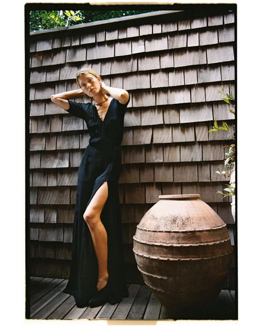 A woman in a black dress poses barefoot on a deck next to a large terracotta pot, set against a weathered cedar shingle wall.