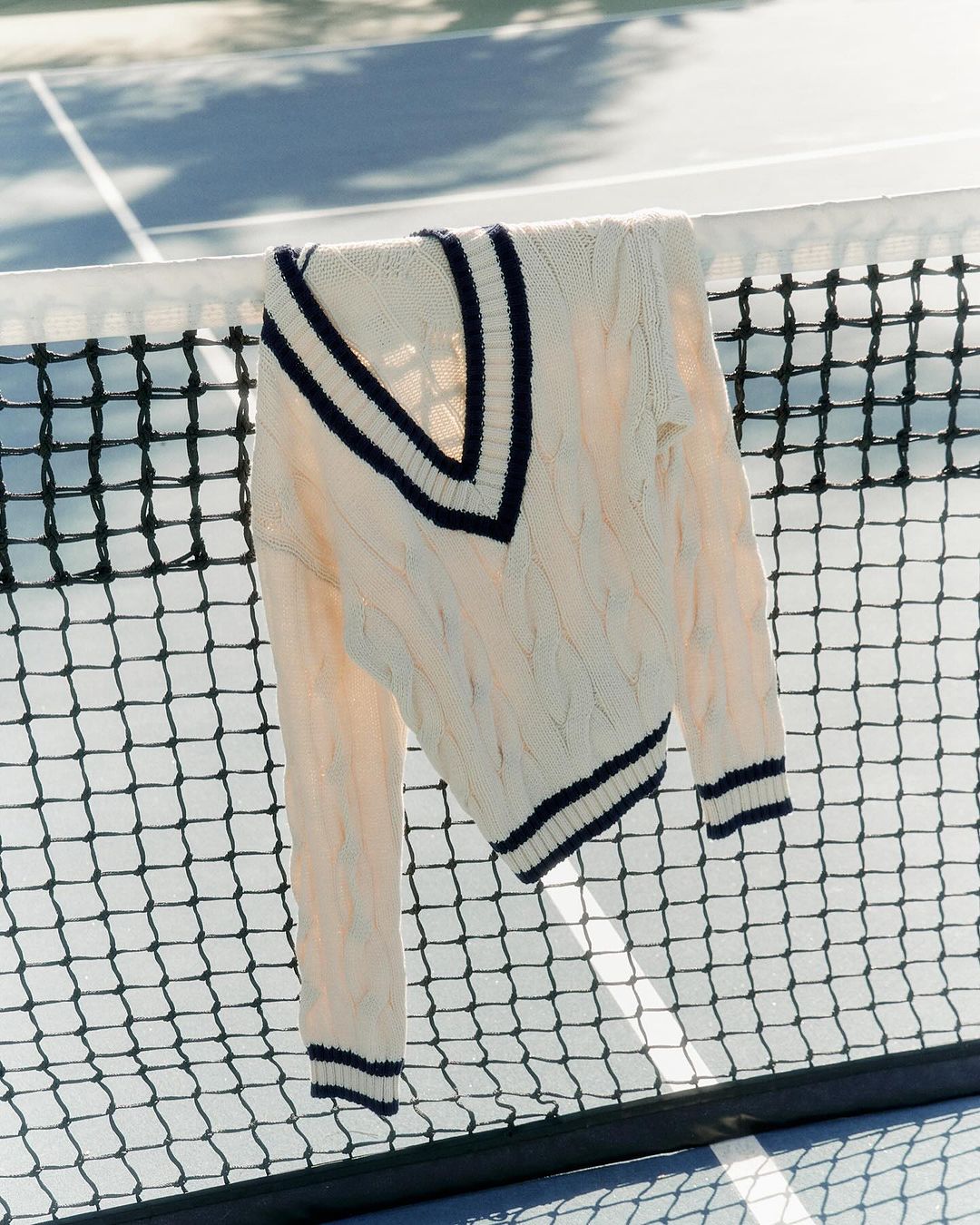 Cable-knit cricket sweater with navy trim hanging on a black tennis net against a blue and white hard court on a sunny day.