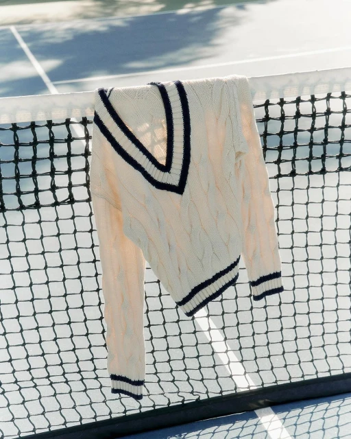 Cable-knit cricket sweater with navy trim hanging on a black tennis net against a blue and white hard court on a sunny day.