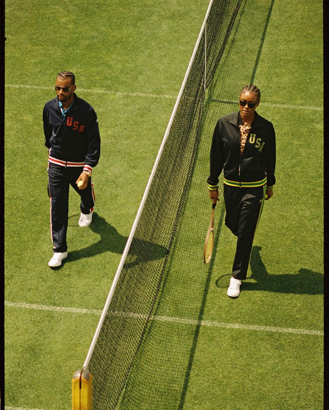 Two athletes in USA tracksuits walk on a tennis court next to the net. One carries a tennis ball and the other a tennis racket.