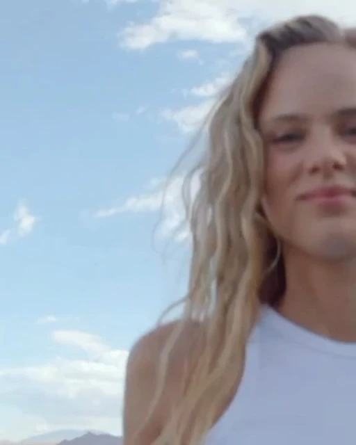 Close-up of a blonde adult female smiling against a sunny sky with fluffy white clouds and distant tan mountains.