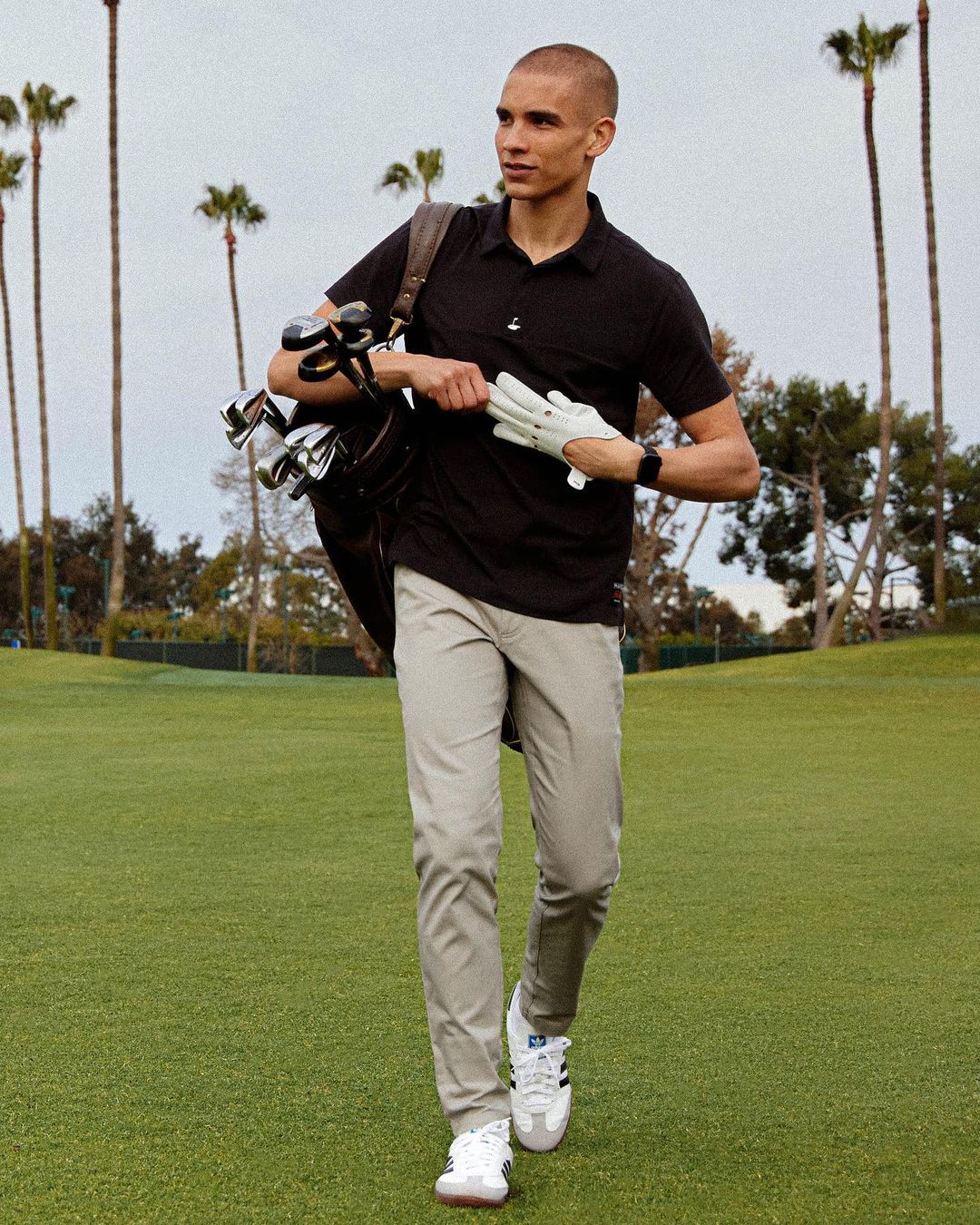 Full shot of a man walking on a golf course, carrying a golf bag and gloves, wearing a black polo shirt and khaki pants.
