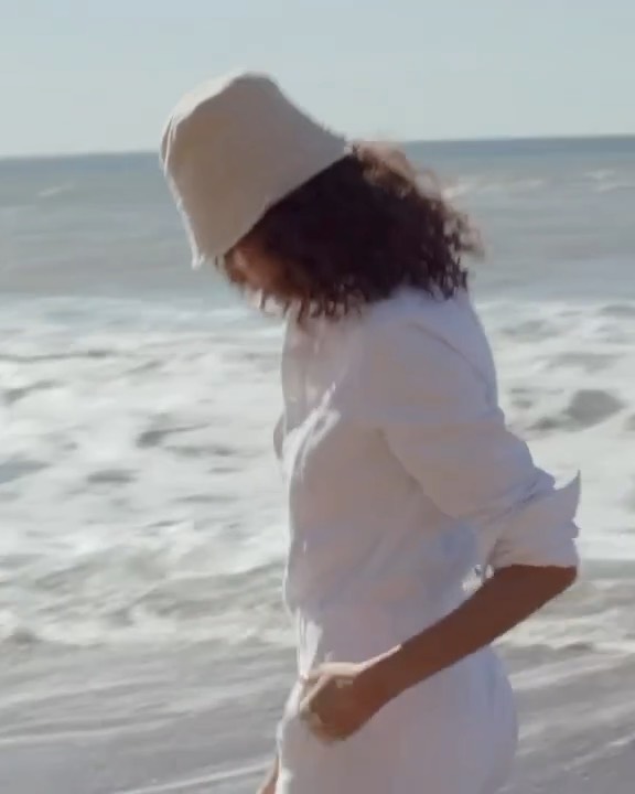 A woman wearing a white bucket hat and white button-down shirt walking along a sandy beach near the ocean.