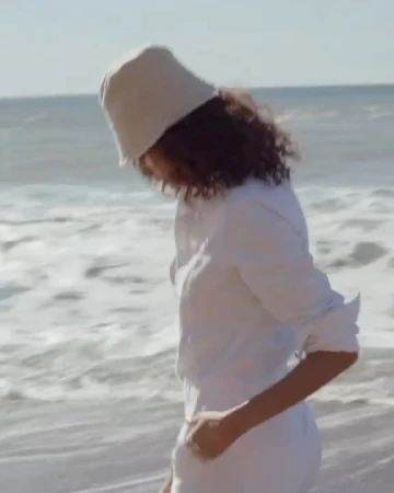 A woman wearing a white bucket hat and white button-down shirt walking along a sandy beach near the ocean.