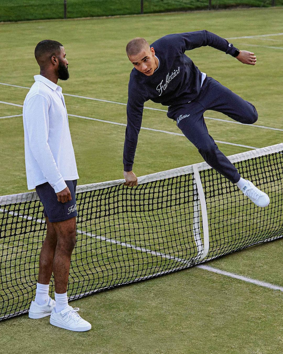 Two men on a tennis court, one in a white polo and navy shorts, and the other leaping over the net in a navy sweatshirt and sweatpants.