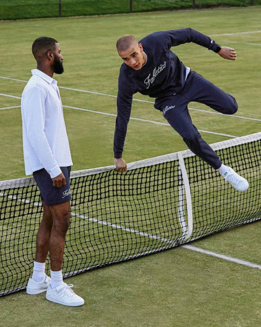 Two men on a tennis court, one in a white polo and navy shorts, and the other leaping over the net in a navy sweatshirt and sweatpants.