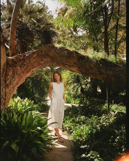 Woman in white dress poses on stone path surrounded by green foliage and a tree archway in an outdoor setting.