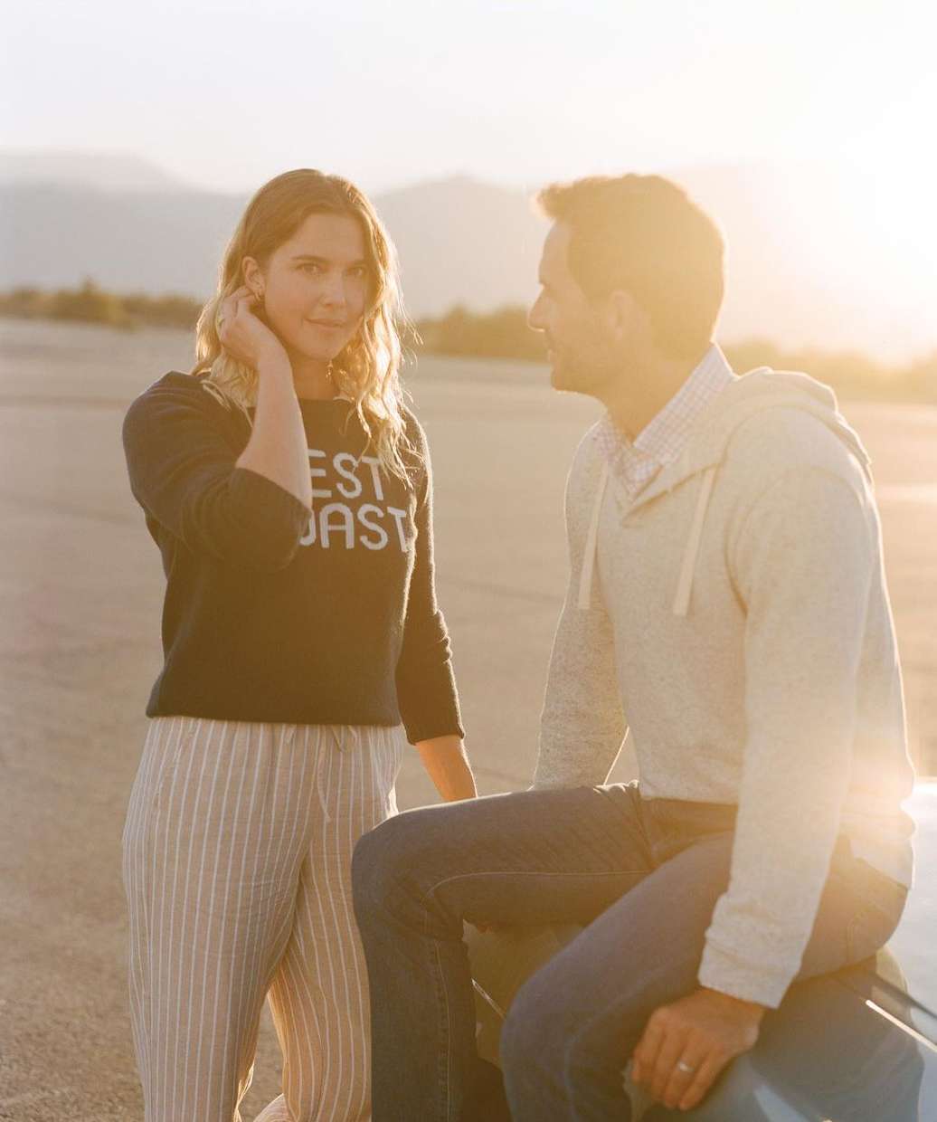 A happy couple stands in a desert landscape at sunset; the woman wears a sweater and striped pants, while the man wears jeans.