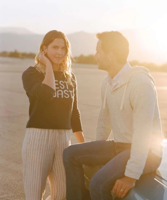 A happy couple stands in a desert landscape at sunset; the woman wears a sweater and striped pants, while the man wears jeans.