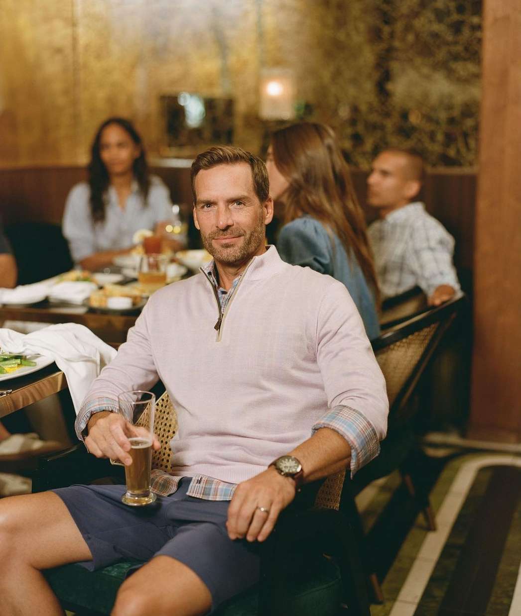 Man in pink sweater sits with beer at a restaurant, surrounded by other people and tables with food and drinks. Warm lighting.