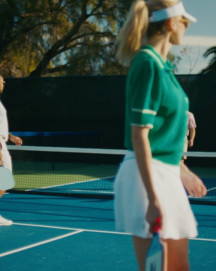 People playing pickleball on an outdoor blue court, wearing athletic clothing and visors, with trees in the background on a sunny day.