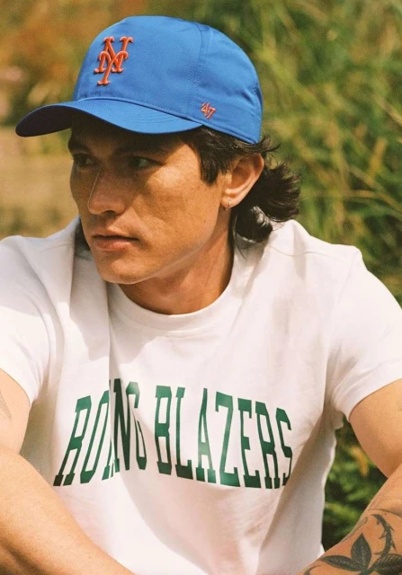 Man wearing baseball cap and t-shirt poses outdoors in bright daylight. He has dark hair, medium skin tone, and a relaxed facial expression.