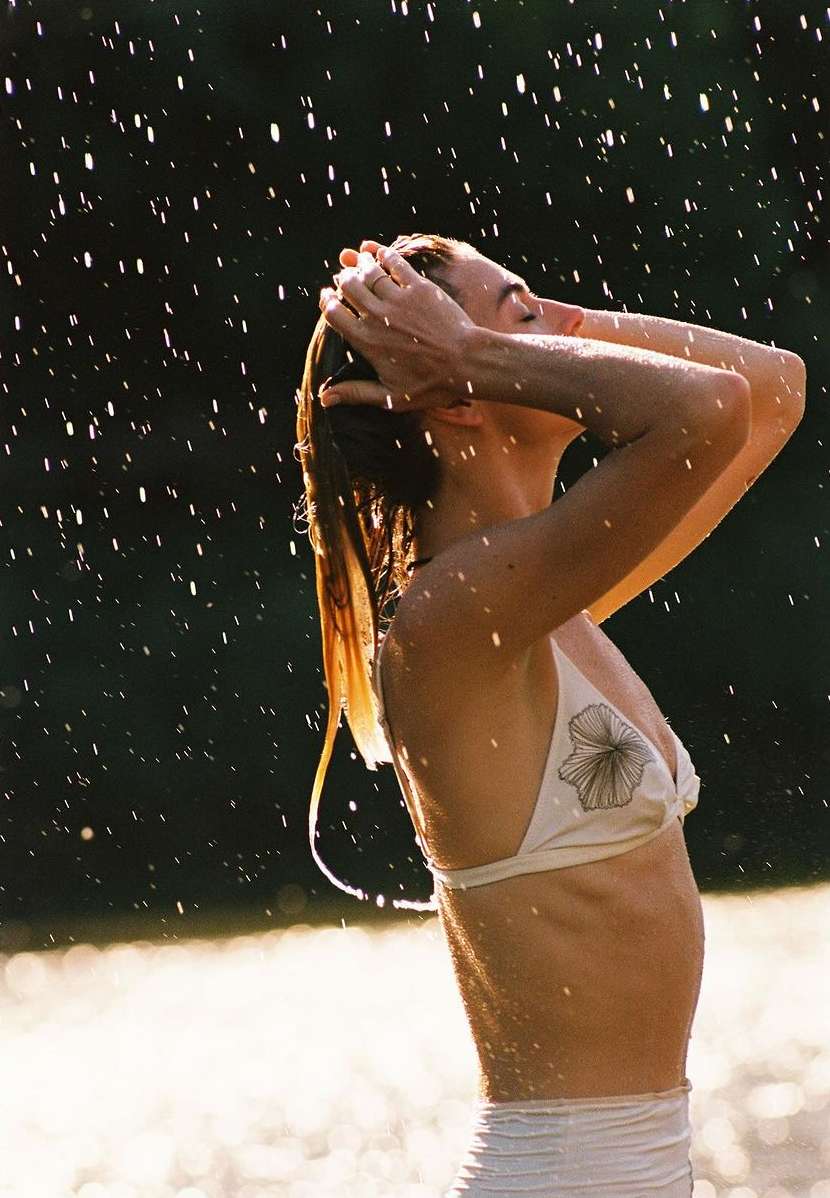 Woman in a bikini and bottoms stands under the rain in an outdoor setting, with hands in her hair and dark blurred background.