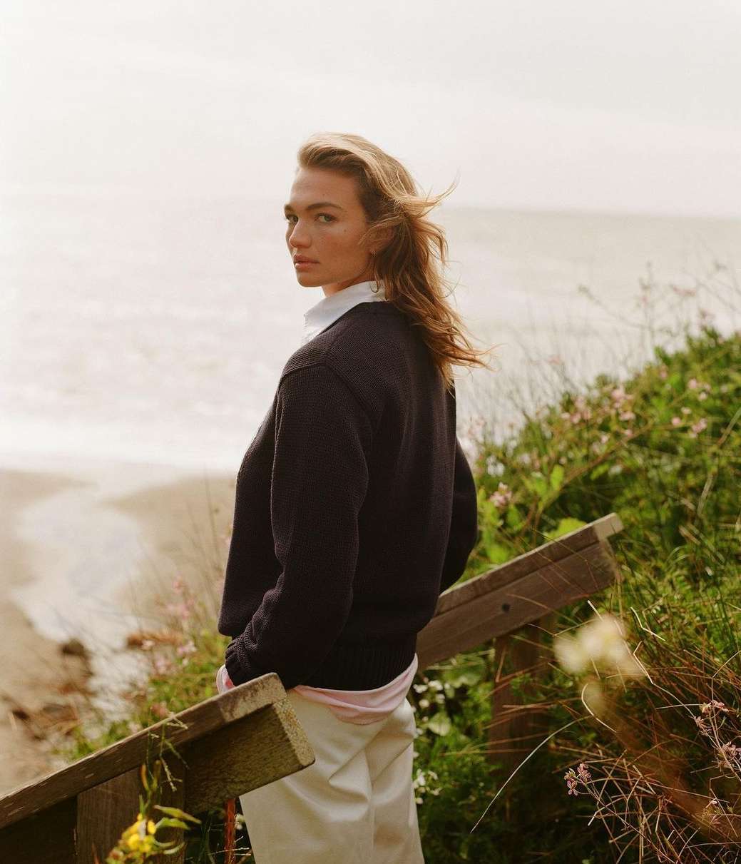 Woman wearing a navy sweater standing near the ocean on a cloudy day. The model looks over her shoulder towards the camera.