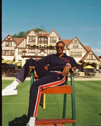 Black woman wearing a navy tracksuit with red piping seated on a tall wooden umpire chair outdoors on a green lawn.